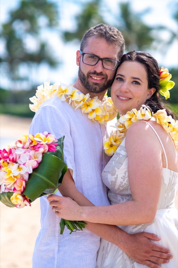 Smiling couple wearing yellow and white plumeria leis, the bride holding a tropical bouquet