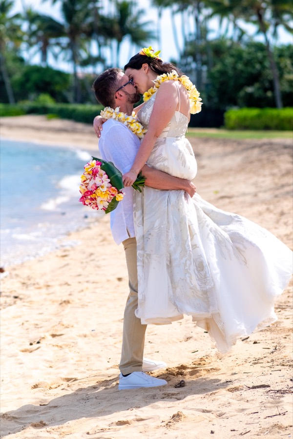 Groom lifting his bride for a kiss on the beach, her gown flowing in the breeze