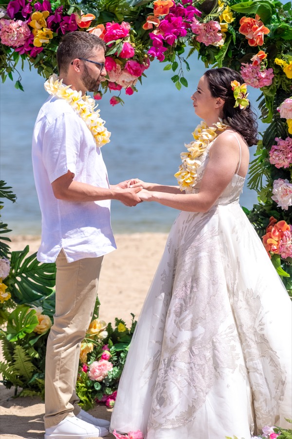 Bride and groom in plumeria leis holding hands during their ceremony under the flower arch