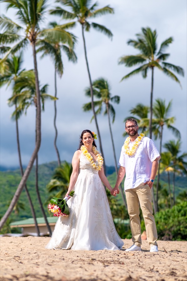Bride and groom holding hands on the sand beneath tall palm trees and green mountains