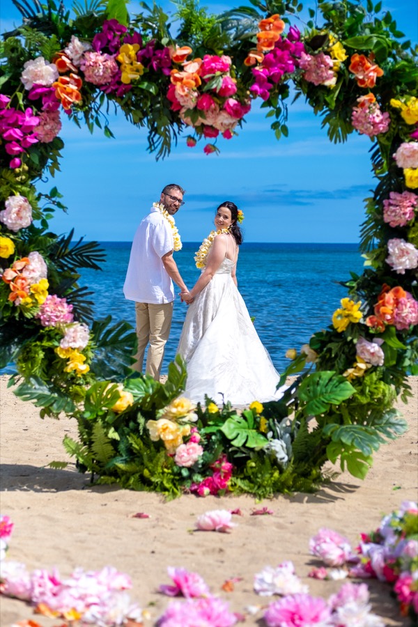 Bride and groom holding hands inside a round tropical flower arch on the beach, with pink petals on the sand