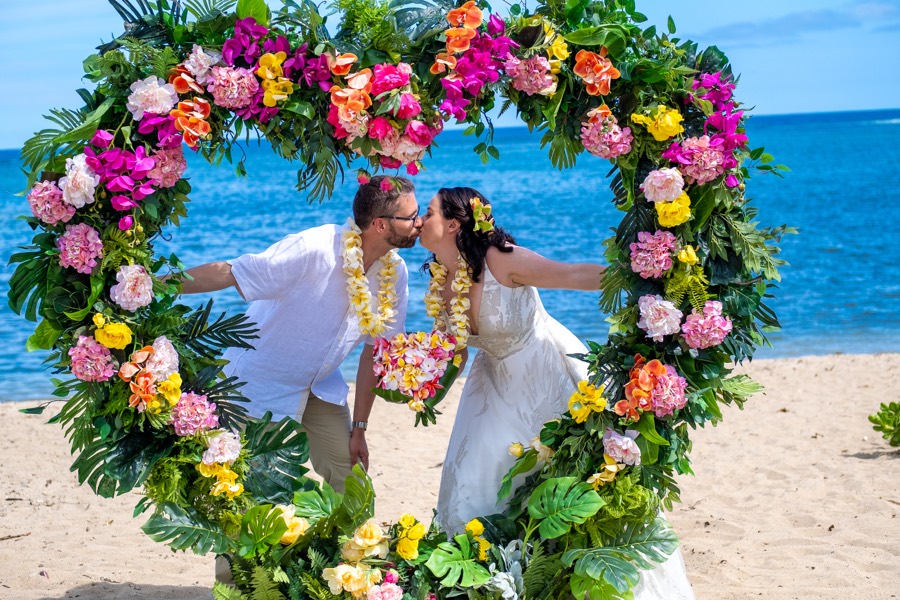 Couple kissing in the middle of a round arch of pink, orange and yellow tropical flowers by the ocean