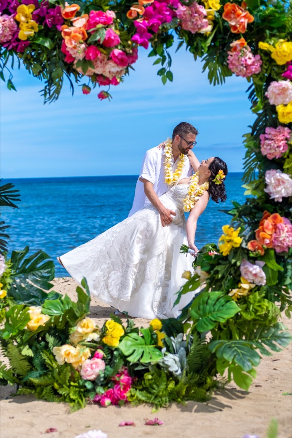 Groom dipping his bride under the tropical flower arch with the blue ocean behind them
