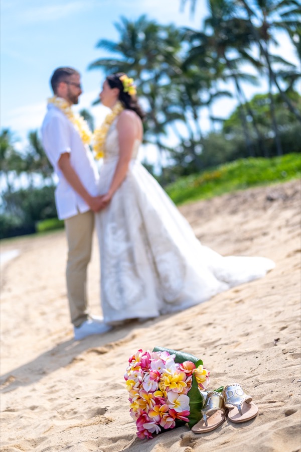 Tropical plumeria bouquet and the bride's sandals on the sand as the couple embraces behind