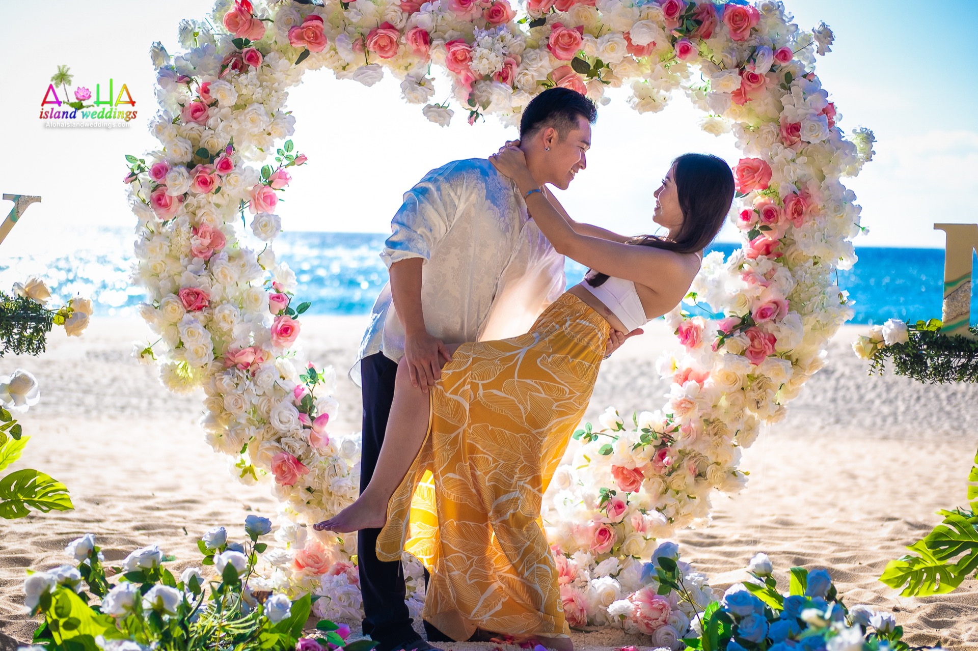 Newly engaged couple dipping in front of a pink rose and white hydrangea heart on the beach