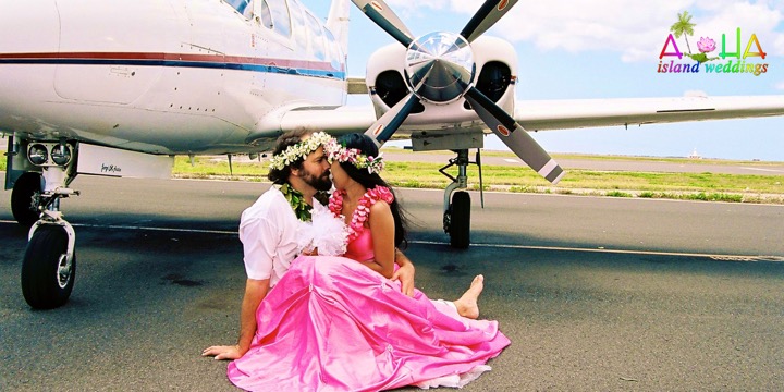 Paul and Dewi in haku leis sitting together beside a small airplane, Dewi in a bright pink gown