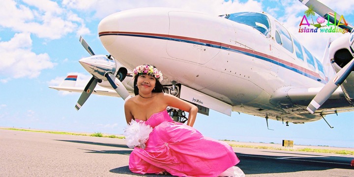 Dewi in a pink gown and flower haku lei posing beside a white airplane
