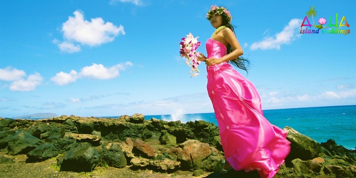 Dewi in a pink gown holding her bouquet on the lava rocks by the ocean