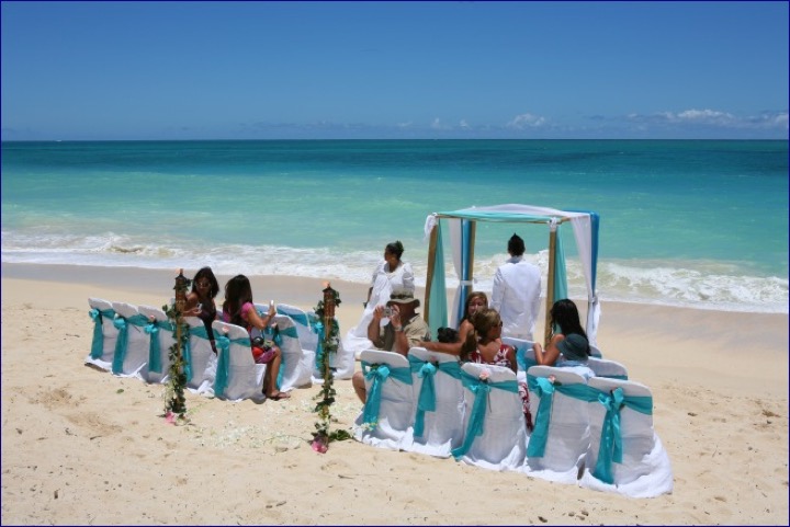 Guests seated on chairs with turquoise sashes facing the arch on the beach