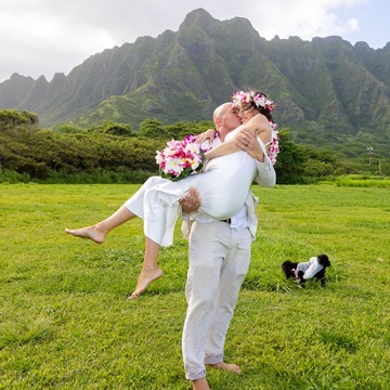 Groom carrying his bride on the grass below Kualoa's green mountains