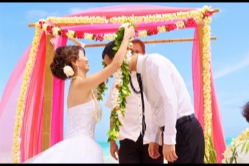 Rei places a maile lei on her groom under a pink flower arch