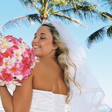 A smiling bride with a pink plumeria bouquet under the palms at Magic Island