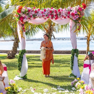 Officiant under a pink and white flower arch on an oceanfront park lawn