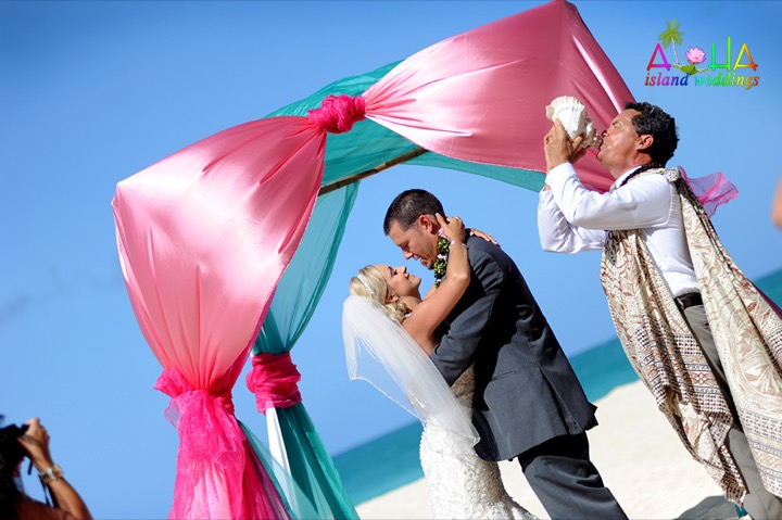 Rev. Kimo sounding the conch shell under a pink draped arch as the couple kisses