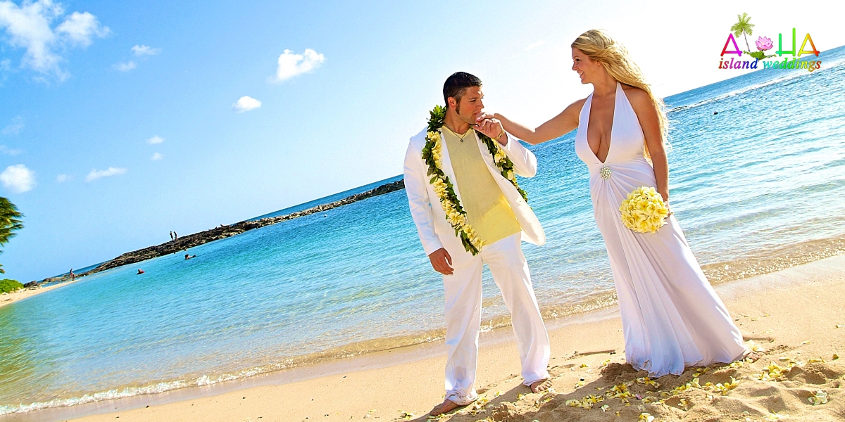 The groom wears a Maile lei entwined with yellow plumeria flowers matching his brides bouquet , he kisses her hand gently by the side of the waters