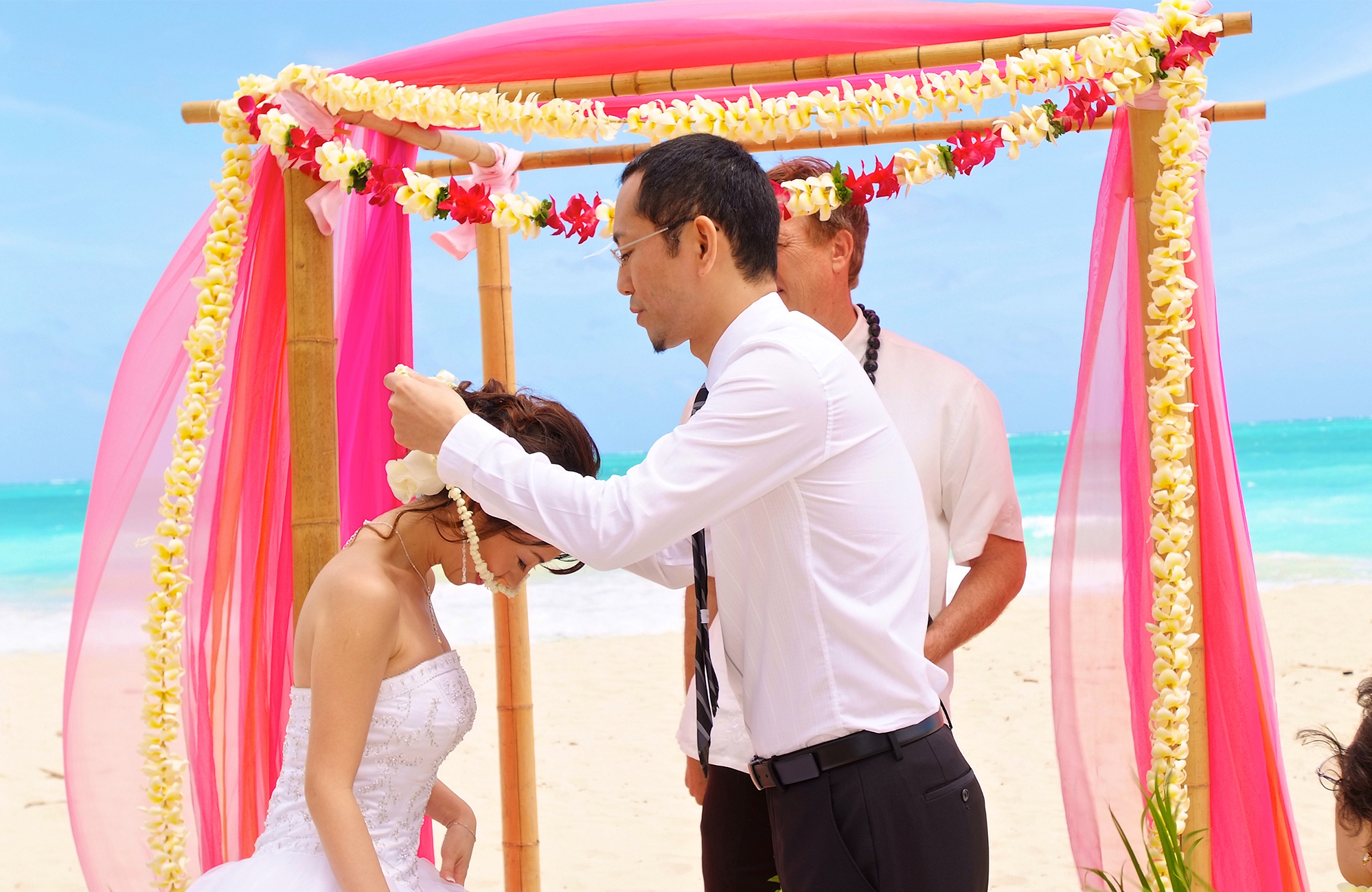 Makoto places a flower lei on his bride Rei under a pink flower arch on Oahu