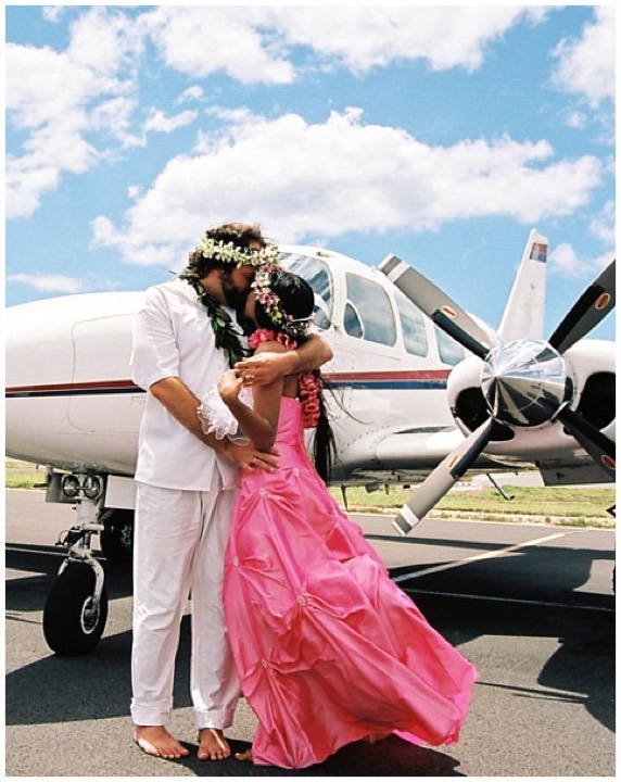Paul and Dewi embracing by an airplane, both wearing haku leis