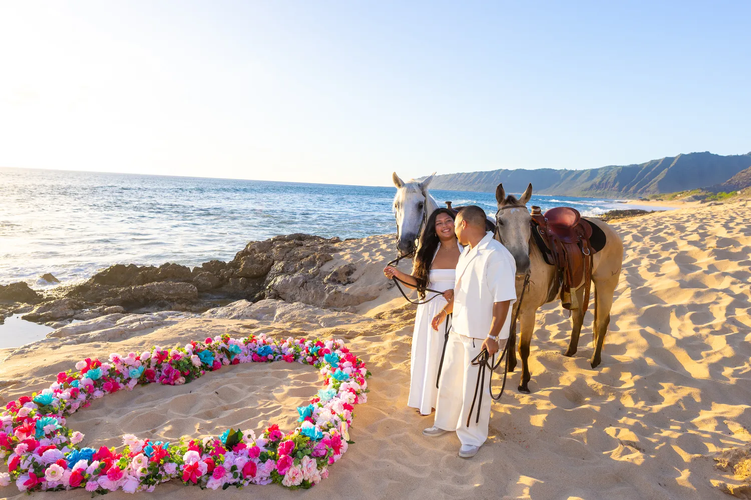 Couple with white and buckskin horses by the ocean, at a horseback proposal on Oahu