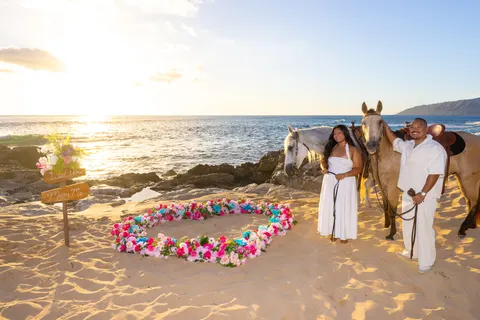 Couple with two horses by flower heart at golden hour, at a horseback proposal on Oahu