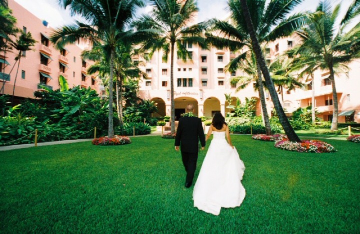Bride and groom walking across the lawn of the pink Royal Hawaiian Hotel under the palms