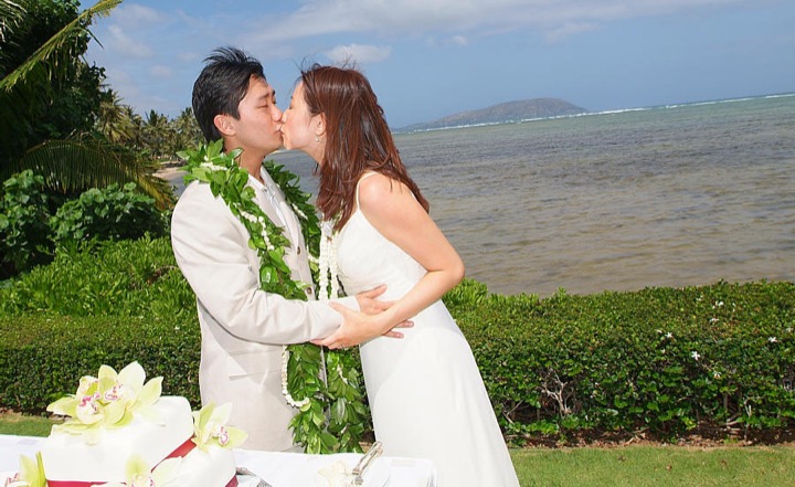 Newlyweds kissing on the lawn by the ocean at the Kahala Resort