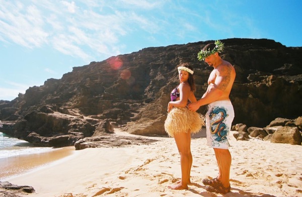 Jamillah and Brandon in grass skirts and haku leis at the cove, from a distance