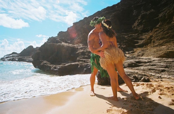 Jamillah and Brandon in grass skirts and haku leis at the cove, smiling
