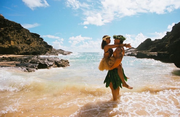 Jamillah and Brandon in grass skirts and haku leis at the cove, on the sand