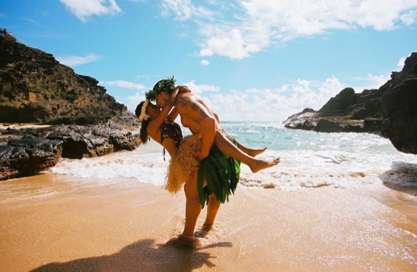 Jamillah and Brandon in grass skirts and haku leis at the cove, looking at each other