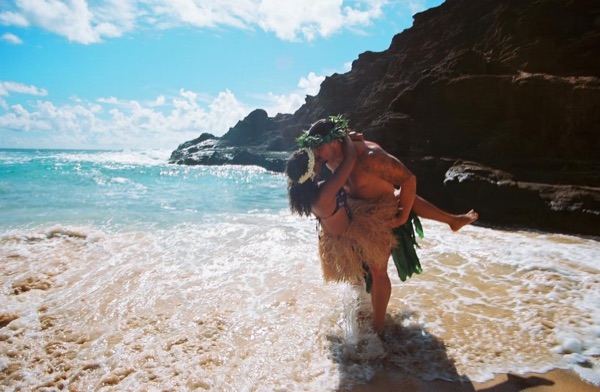 Jamillah and Brandon in grass skirts and haku leis at the cove, wide view