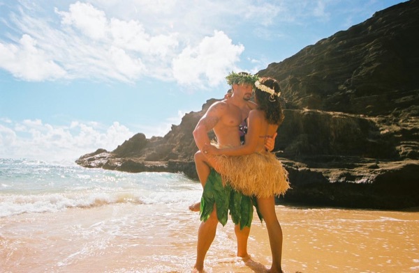 Jamillah and Brandon in grass skirts and haku leis at the cove, under a blue sky