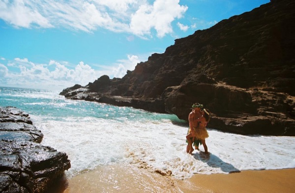Jamillah and Brandon in grass skirts and haku leis at the cove, in the sunshine