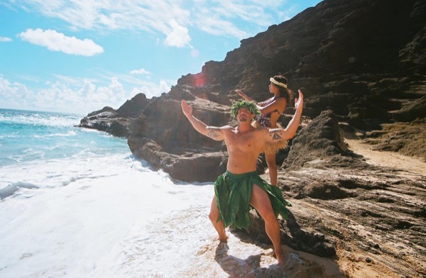 Jamillah and Brandon in grass skirts and haku leis at the cove, in a playful moment