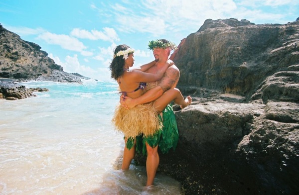 Jamillah and Brandon in grass skirts and haku leis at the cove, with a big smile