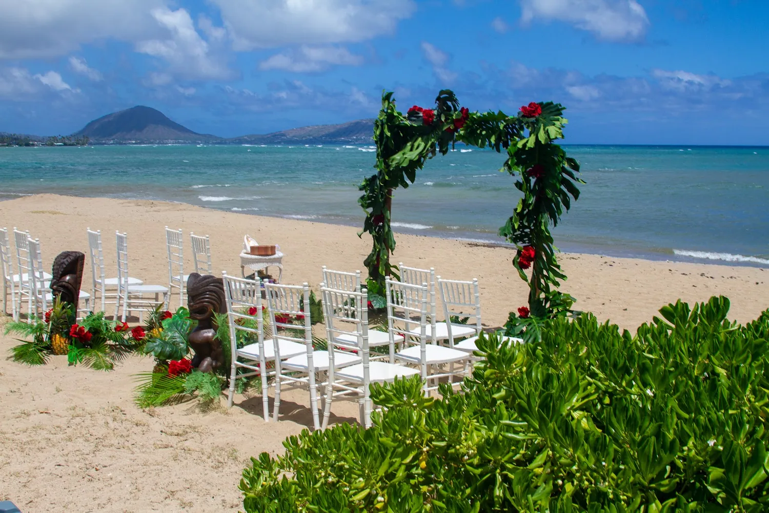 A green leaf arch with red flowers and white chairs by the ocean
