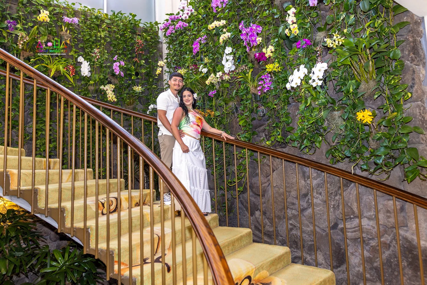 The couple on a spiral staircase beside a wall of orchids