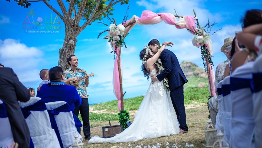 Bride and groom kiss under the pink draped branch arch as the ukulele player plays and guests watch