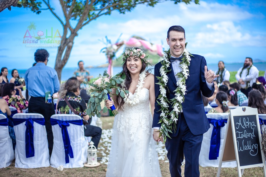 Smiling newlyweds in white plumeria leis walk back down the aisle past chairs with blue sashes