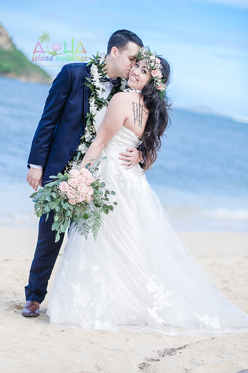 Groom kisses his bride's cheek on the beach at Kualoa, with Chinaman's Hat in the distance
