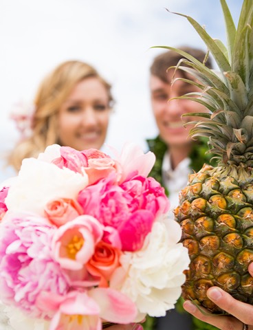 A pink peony bouquet and a pineapple, with the groom in a maile lei behind them in Hawaii