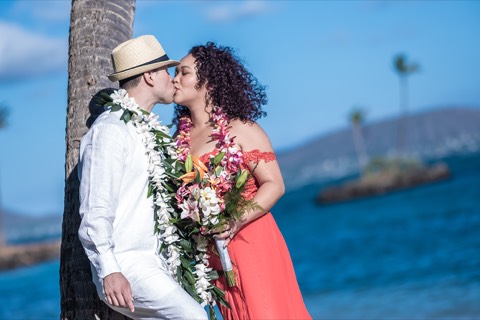 Maya and her groom kiss against a palm tree in their flower leis at Waialae Beach Park