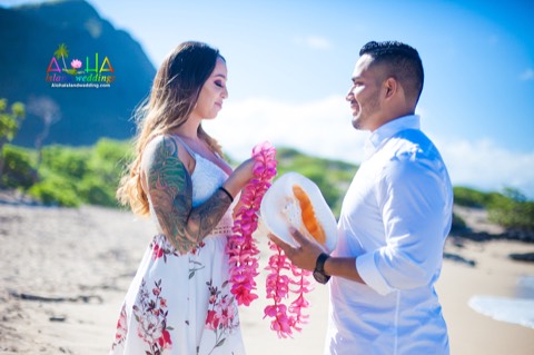 A bride gives her groom a plumeria flower lei as he holds a conch shell at Makapuu beach on Oahu
