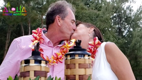 A vow renewal couple in orange flower leis kissing behind two lit tiki torches in Hawaii