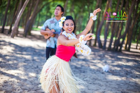 A smiling hula dancer in a grass skirt and white flower leis dancing under the ironwood trees on Oahu