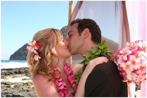 Tara and her groom kiss at Makapuu beach in pink and chocolate plumeria leis