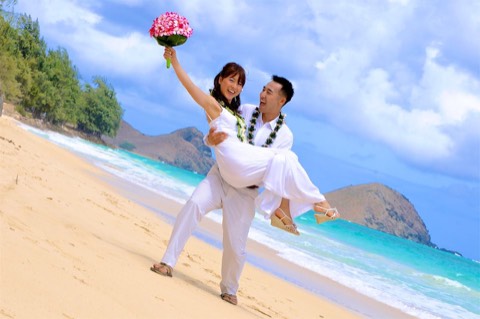 A groom in a maile lei lifts his bride with her plumeria bouquet on the beach in Hawaii