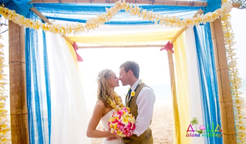 A kiss under a bamboo arch strung with yellow plumeria leis, the bride in a plumeria lei on Oahu