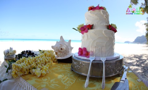 A white wedding cake with flower leis and a conch shell by the ocean in Hawaii