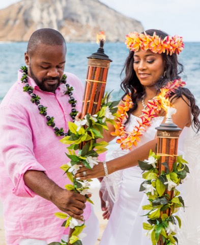 A couple in flower leis lighting bamboo tiki torches together as a unity ceremony on Oahu