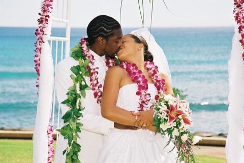 A couple kisses in orchid leis under white drapes by the ocean on Oahu